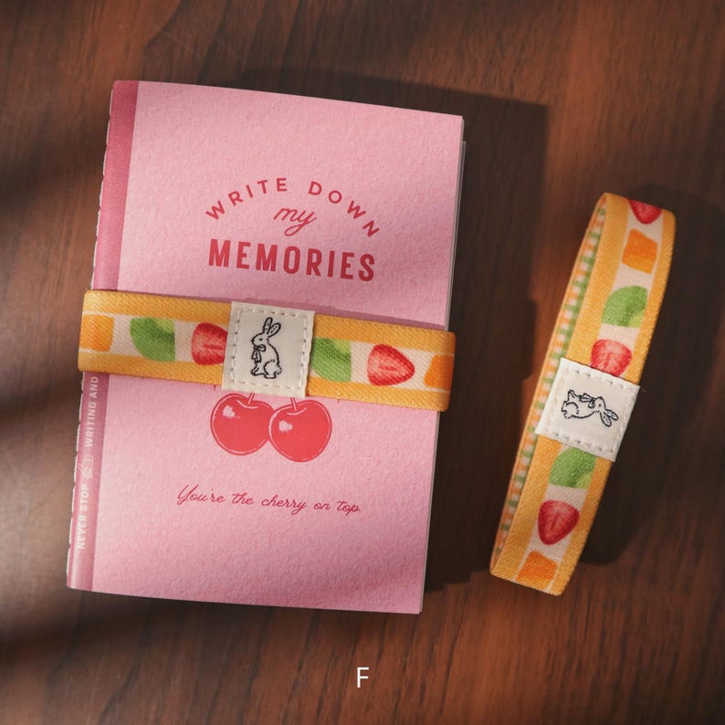 May include: A pink notebook with the text "Write Down My Memories" and "You're the cherry on top." A yellow elastic band with fruit designs and a bunny logo wraps around the notebook. A matching band lies next to it.