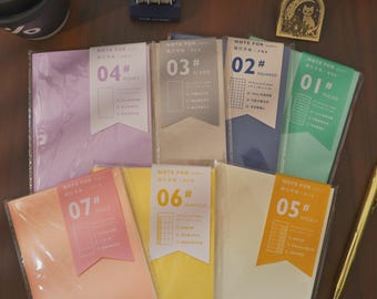 May include: A collection of colorful notebooks with different page styles, including ruled, squared, and blank. Each notebook has a unique color and a label indicating its type, such as "Daily" or "Weekly". A gold pen and other stationery items are also visible.