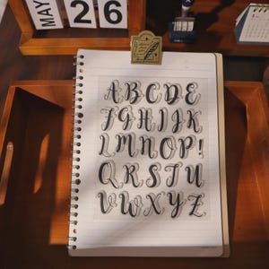 May include: A clear plastic sheet featuring the full alphabet in a black cursive script font, arranged in five rows on a white spiral-bound notebook. The letters are printed in a calligraphy style with a distinct shadow effect behind each character.
