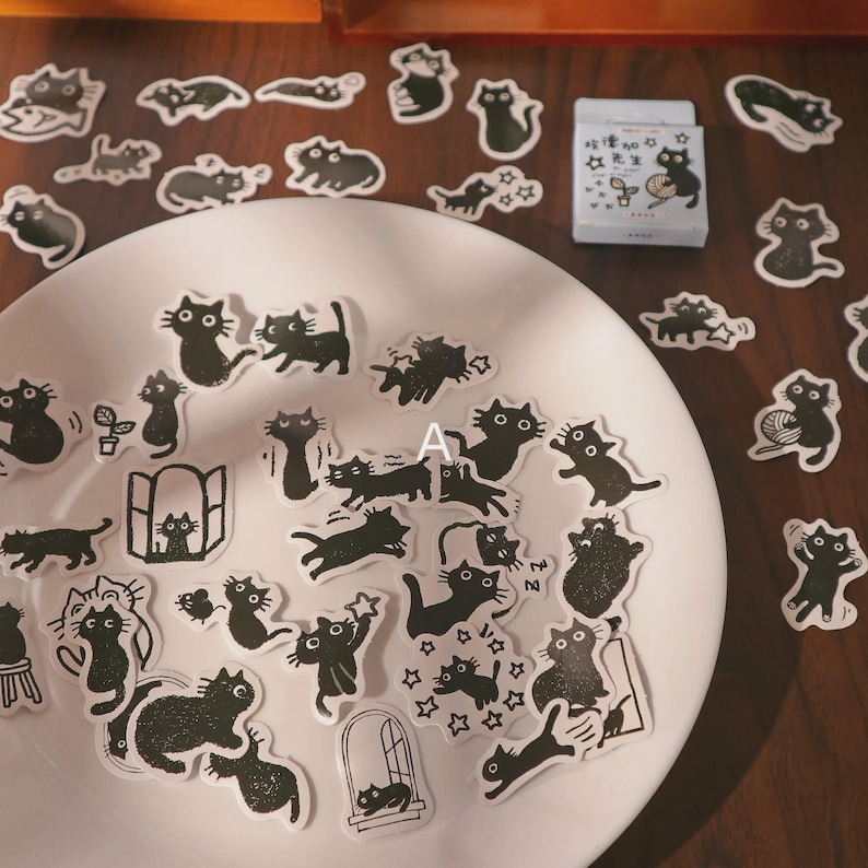 May include: A collection of black cat stickers in various poses, including cats with stars, plants, and yarn. The stickers are arranged on a white plate and a wooden surface. A small box with a cat illustration is also present.