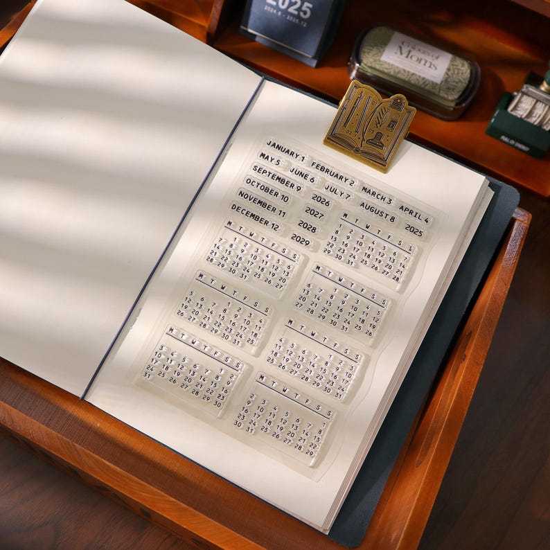 May include: An open notebook with a clear calendar stamp set. The stamp set includes monthly calendars for the years 2025-2029. A gold-colored metal bookmark shaped like a book is also visible.