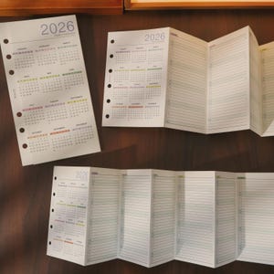 May include: White 2026 yearly overview calendars with monthly layouts and lined pages. The calendars have a hole-punched edge for binding. The months are colour-coded. The calendars are on a dark brown surface.