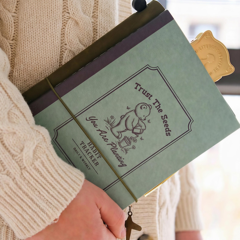 May include: A green habit tracker notebook with the text "Trust The Seeds You Are Planting." The cover features a bear watering a plant. A gold-colored bookmark and a brown elastic band secure the notebook. The notebook is held by a person wearing a cream-colored sweater.