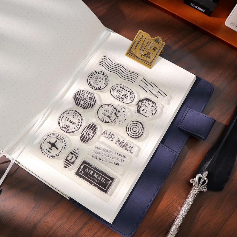 May include: Clear stamp set with various vintage-style postage designs, including "Air Mail" text, airplane, and city names. The stamps are on a clear sheet, resting on an open notebook with a blue cover. A gold-colored book-shaped paper clip is also present.