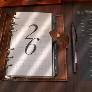 May include: Open brown leather planner with a 2026 calendar page visible. The calendar page has large numbers '26' and monthly grids. Black divider tabs read 'Finances', 'Notes', 'Habits', 'Home', and 'Personal'. A brown pen rests beside the planner.