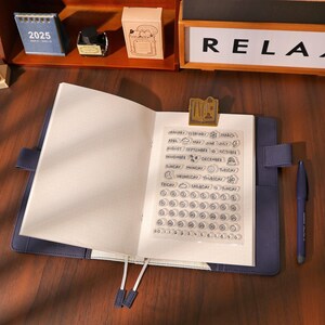 May include: Open blue journal with a clear stamp sheet featuring months, days, and numbers. A gold-colored clip holds the stamp sheet in place. A blue pen rests on the right side. A small 2025 calendar and other stationery items are in the background.