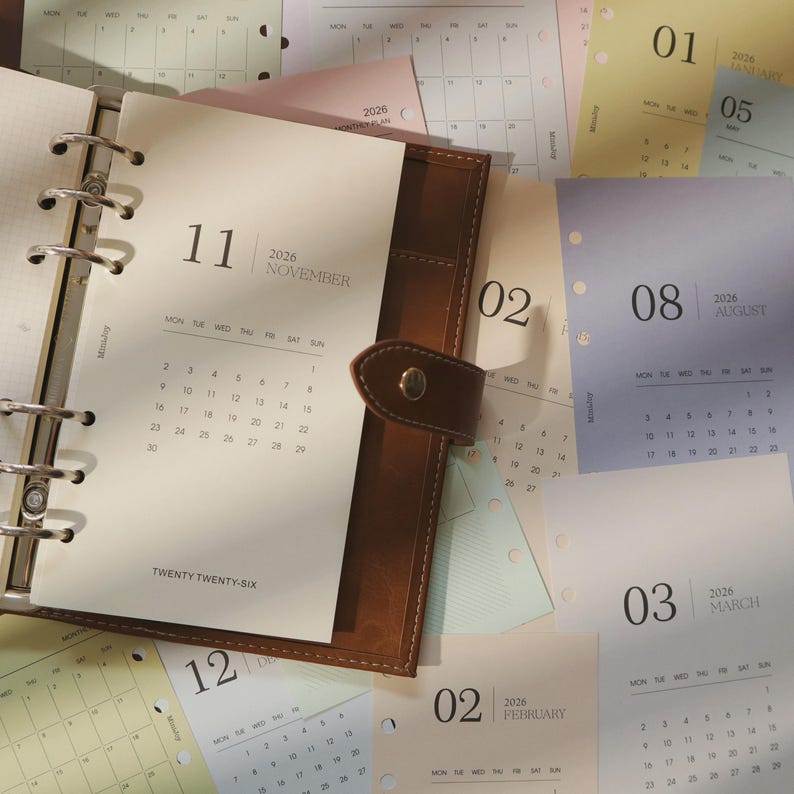 May include: A brown leather planner open to November 2026, with a metal binder and snap closure. Surrounding the planner are loose-leaf calendar pages for different months, including January, February, March, and August.