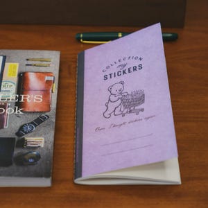 May include: A light purple sticker collection notebook featuring a black line drawing of a teddy bear pushing a shopping cart filled with stickers. The cover text reads COLLECTION my STICKERS and Oops. I bought stickers again. A dark pen lies above.