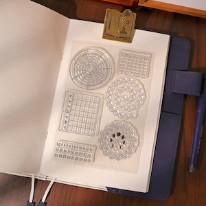May include: Clear acrylic stamps with various calendar and circular designs, including a monthly calendar, a weekly calendar, and a lunar cycle. The stamps are on a white sheet of paper inside a blue journal.