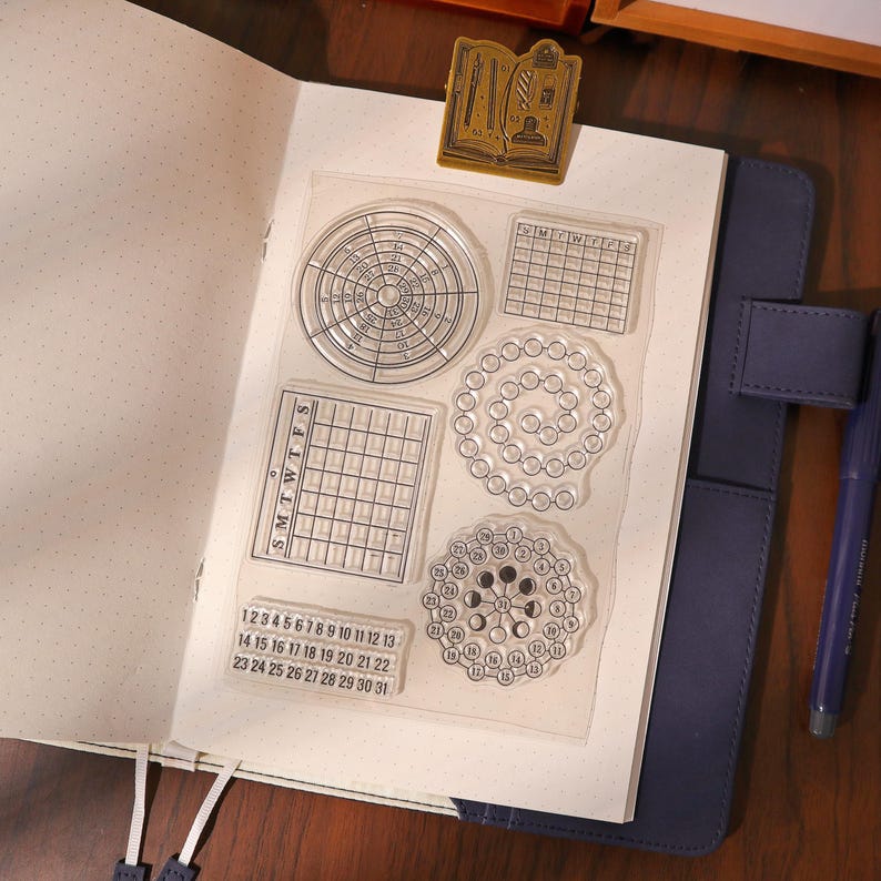 May include: Clear acrylic stamps with various calendar and circular designs, including a monthly calendar, a weekly calendar, and a lunar cycle. The stamps are on a white sheet of paper inside a blue journal.