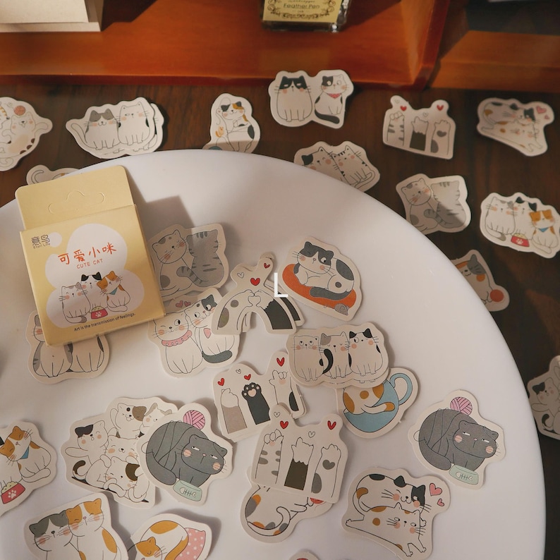May include: A collection of cat-themed stickers scattered on a white plate. The stickers feature various cat illustrations in different poses and colors, with some holding hearts. A small box with Chinese text is also present, labeled "Cute Cat".