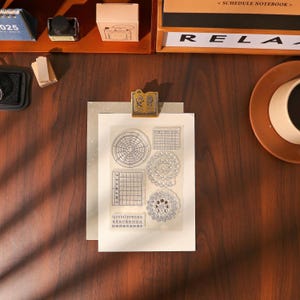 May include: A clear stamp set with various calendar and circular designs, held by a gold-colored clip. The stamp set includes a monthly calendar, a weekly calendar, and a numbered date grid. The background includes a wooden desk and stationery.
