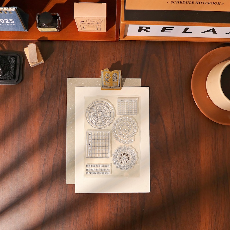 May include: A clear stamp set with various calendar and circular designs, held by a gold-colored clip. The stamp set includes a monthly calendar, a weekly calendar, and a numbered date grid. The background includes a wooden desk and stationery.
