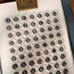 Pode incluir: Carimbos circulares transparentes com letras, números e símbolos pretos. As letras de A a Z, os números de 0 a 9 e símbolos como o & e o ponto de interrogação são visíveis. Um carimbo de madeira está no canto superior esquerdo.