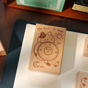 May include: Wooden stamp with a cat design and the text "What Should I Do Today?" The stamp includes a clock face with "Date" in the center, a sun, a moon, and paw prints. The stamp is made of light-colored wood.