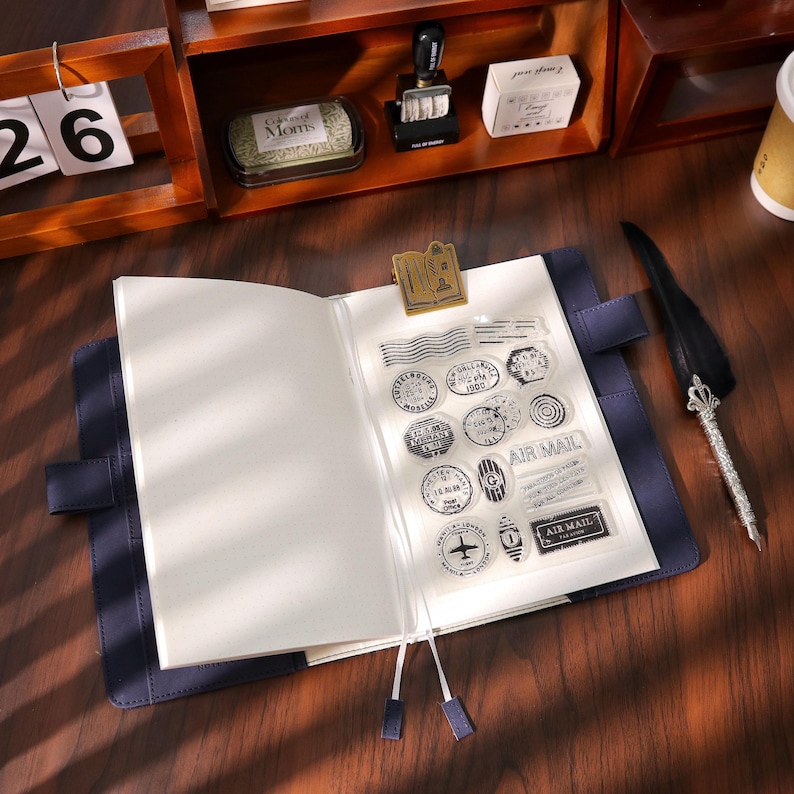 May include: An open blue leather journal with blank pages, held open by a gold-colored metal clip. Clear stamps with various circular and rectangular designs are visible. A decorative pen and a wooden shelf with stationery items are in the background.