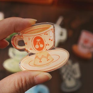 May include: A clear acrylic charm depicting a cup of coffee with a cherry design and a dessert on a saucer. The cup and saucer are light orange with red cherry accents. The charm is held in a person's hand.