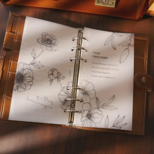 Peut inclure: Un agenda en cuir ouvert, avec des illustrations florales sur les pages. Les pages présentent une écriture manuscrite et le texte "YOUR OWN STORY". L'agenda est ouvert sur une surface en bois.