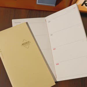 May include: A tan-colored weekly travel notes journal with the text "WEEKLY TRAVEL NOTES" and a weekly planner layout. The open journal shows days of the week, with Thursday, Friday, Saturday, and Sunday visible. The journal is on a wooden surface.