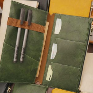 May include: Open green leather journal with two gray pens secured by a brown elastic band. The journal has multiple pockets and a zipper closure. The interior is tan and includes a notepad. Other journals in various colors are visible in the background.
