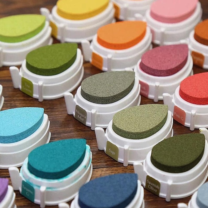 May include: A collection of colorful ink pads in various shades, including green, blue, pink, and orange. Each ink pad has a teardrop-shaped sponge and a white plastic base. Some pads have labels with names like "Niagara Mist" and "Sage".