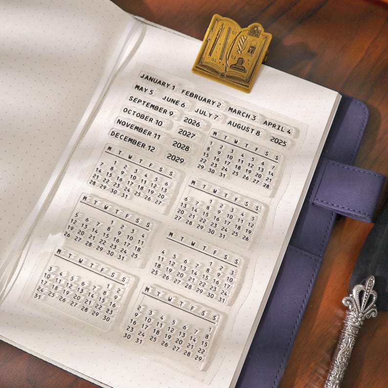 May include: Clear acrylic calendar stamps with dates from 2025 to 2029, displayed on a dotted paper notebook. A gold-colored book-shaped paper clip is attached to the notebook. The stamps are arranged to show the months and days.