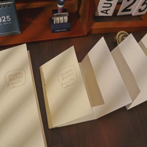 May include: Cream-colored travel notebooks with the text "TRAVEL NOTES freewriter" in a handwritten style. The notebooks are open, revealing blank pages, and are arranged on a dark wooden surface. A calendar and other stationery items are in the background.
