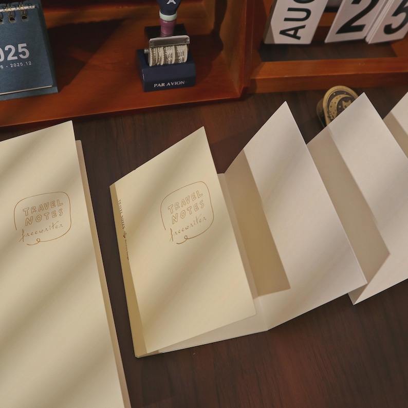 May include: Cream-colored travel notebooks with the text "TRAVEL NOTES freewriter" in a handwritten style. The notebooks are open, revealing blank pages, and are arranged on a dark wooden surface. A calendar and other stationery items are in the background.
