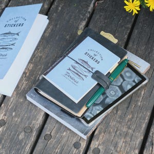 May include: A stack of notebooks and a tablet rest on a wooden surface. The top notebook has a light blue cover with the text "COLLECTION of STICKERS" and illustrations of sardines. A green pen is clipped to the stack.