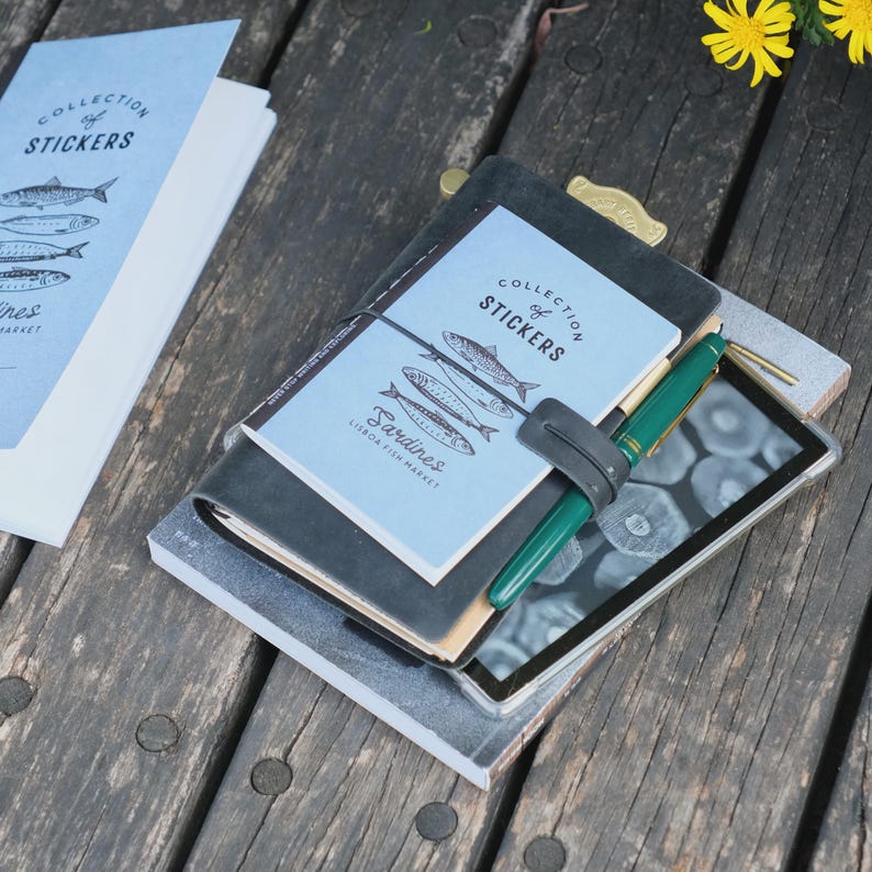 May include: A stack of notebooks and a tablet rest on a wooden surface. The top notebook has a light blue cover with the text "COLLECTION of STICKERS" and illustrations of sardines. A green pen is clipped to the stack.