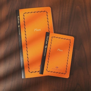 Könnte beinhalten: Zwei orangefarbene Notizbücher mit schwarzen Rücken und dem Wort "Plan" auf der Vorderseite. Die Notizbücher haben einen dekorativen Rand und den Text "Travel notes" und "SENTOSA DESIGN" auf dem Rücken. Die Notizbücher liegen auf einer dunklen Holzoberfläche.