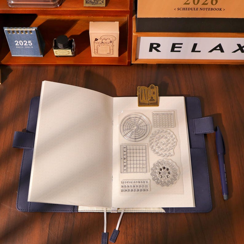 May include: An open blue journal with blank pages, a gold-colored paper clip, and a clear stamp set featuring circular and grid designs. A blue pen rests on the right. Other stationery items are visible in the background.