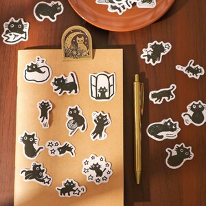 May include: A collection of black and white cat stickers in various poses, including cats with yarn, in windows, and with stars. A gold pen and a brown paper notebook are also visible on a wooden surface. A decorative cat-themed paper clip is attached to the notebook.