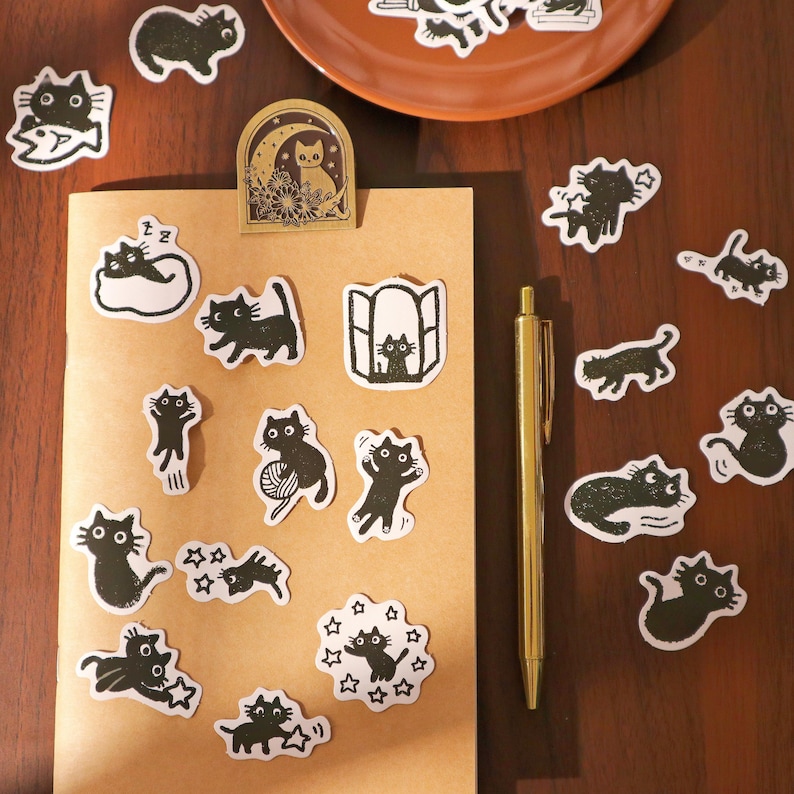 May include: A collection of black and white cat stickers in various poses, including cats with yarn, in windows, and with stars. A gold pen and a brown paper notebook are also visible on a wooden surface. A decorative cat-themed paper clip is attached to the notebook.