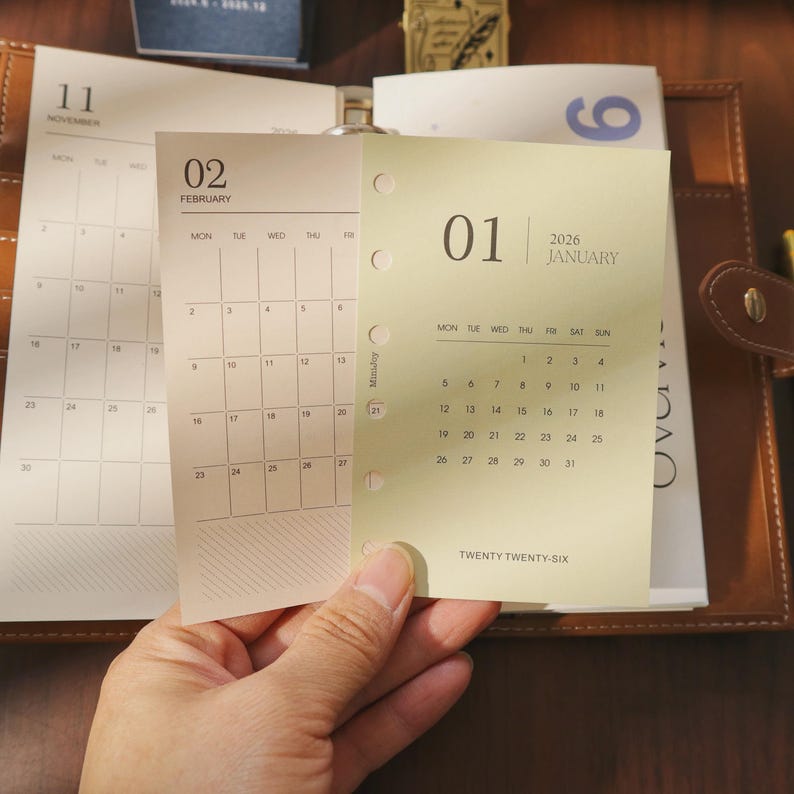May include: A close-up of calendar pages for November, February, and January 2026. The January page is light yellow with the month and year at the top. The calendar pages are held in a brown leather binder.