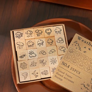 May include: A set of 25 wooden stamps featuring weather-related illustrations, including clouds, rain, sun, and wind. The stamps are arranged in a box with a brown paper label that reads "Weather".