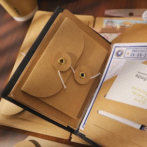 以下が含まれることがあります： 開いた黒革のジャーナル。茶色の紙の封筒とインサートが入っています。封筒は紐と真鍮のハトメで固定されています。ジャーナルには、「IMMIGRATION OFFICE」というテキストと「06-09-2」という日付が印刷されたカードも含まれています。