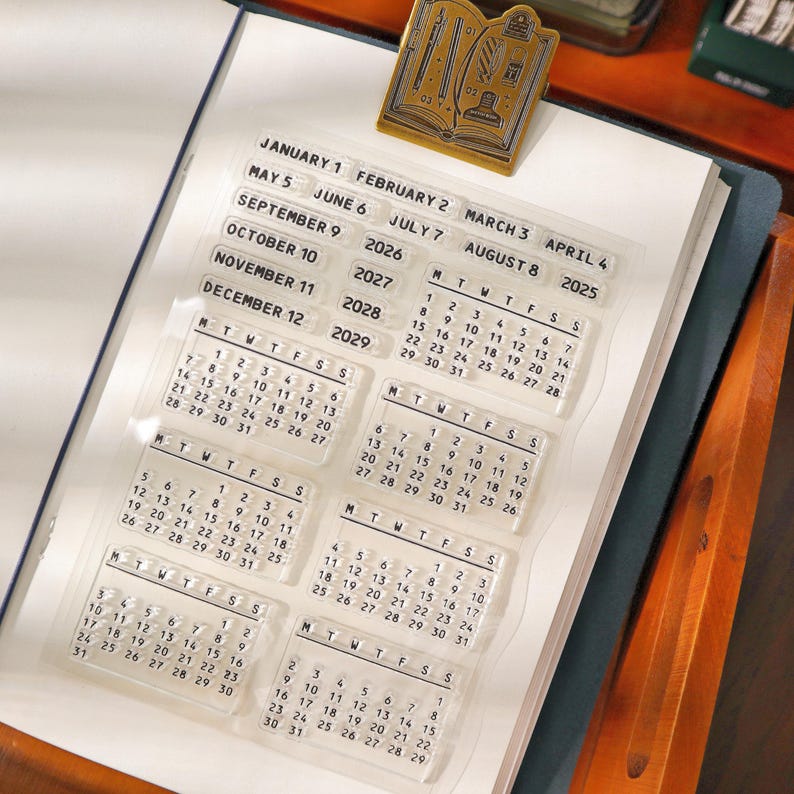 May include: Clear acrylic calendar stamps with monthly and yearly date options, including 2025-2029. A gold-colored metal book-shaped paper clip rests on the open notebook. The stamps are designed for planning and journaling.