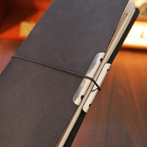 May include: A dark brown leather journal with a metal clip closure and a black elastic band. The journal is open, revealing the cream-colored pages inside. The metal clip is silver.