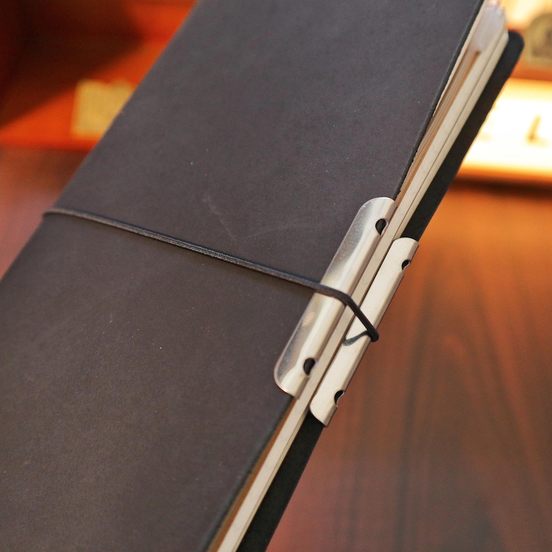 May include: A dark brown leather journal with a metal clip closure and a black elastic band. The journal is open, revealing the cream-colored pages inside. The metal clip is silver.