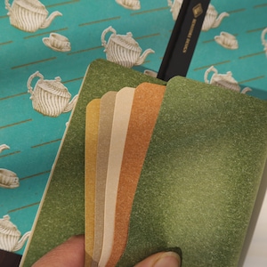 May include: A set of colorful, textured notebooks with a green cover and a variety of interior pages in shades of gold, tan, and orange. The notebooks are held open, revealing the different colored pages. The background features a teal pattern with white teapots.