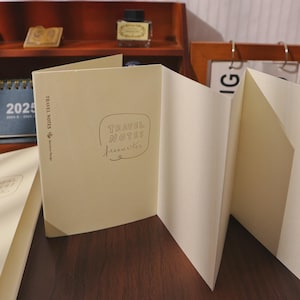 May include: Cream-colored travel journal with the words "TRAVEL NOTES freewriter" in a handwritten style. The journal is open, revealing blank pages. A 2025 calendar and ink bottle are visible in the background.