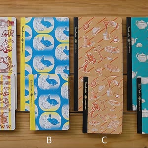 May include: Four notebooks with different cover designs. One has a yellow background with illustrations of bears, cherries, and a rocking horse. Another has a blue background with white rabbits. The third has a tan background with food illustrations. The last has a teal background with teapots.