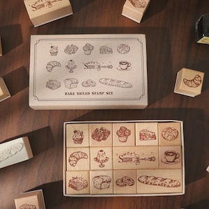 Peut inclure: Un ensemble de tampons Bake Bread dans une boîte en carton rectangulaire avec des illustrations de pâtisseries et une tasse de café. Le set comprend douze tampons en bois individuels aux motifs assortis, dont un croissant, un gâteau et un pain.