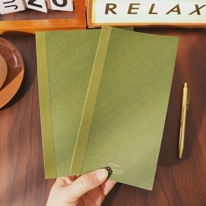 May include: Two olive green notebooks with a matching spine are held in a hand. A gold pen rests on a dark wooden surface next to the notebooks. The word "RELAX" is visible in the background.