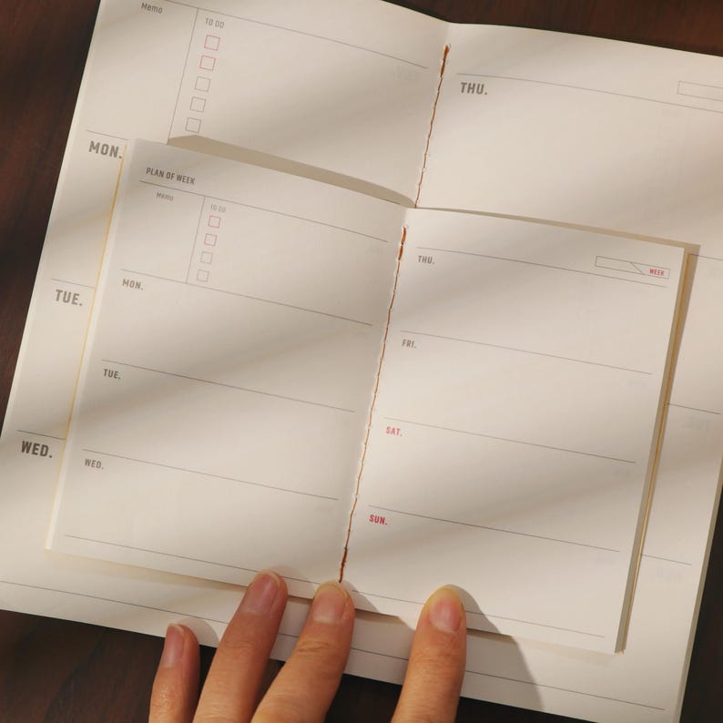 May include: Open weekly planner with a minimalist design. The planner features a cream-colored paper with sections for each day of the week, including a "To Do" list and a "Plan of Week" section. The days of the week are labeled in a simple font.