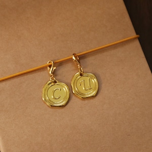 Op de afbeelding: Twee goudkleurige bedeloorbellen. Elke oorbel heeft een cirkelvormig ontwerp met een letter in reliëf in het midden. Eén oorbel heeft de letter "C", de andere de letter "U". De oorbellen zijn opgehangen aan een goudkleurige sluiting.