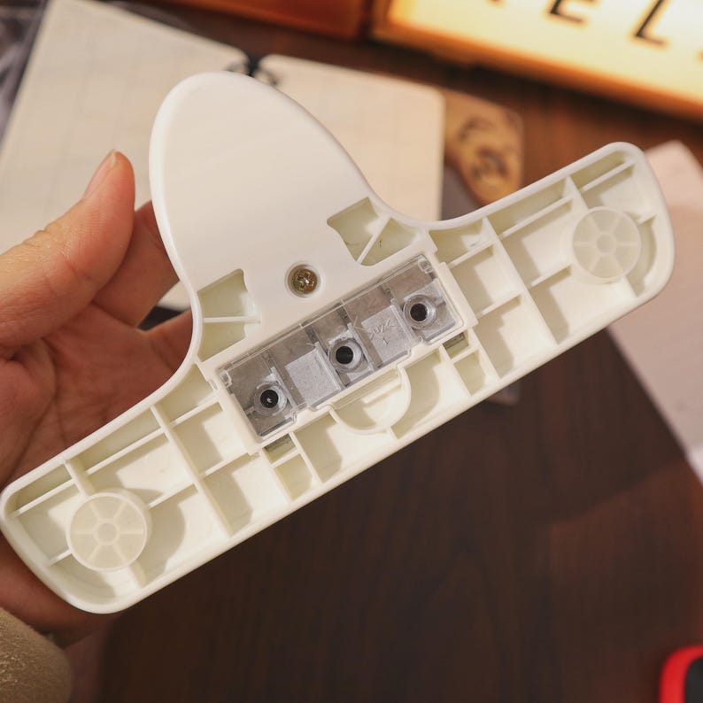 May include: A white plastic hanger with a wide, flat base and a curved top. The underside features a clear plastic section with three small holes. Two circular, ridged details are visible on the base.