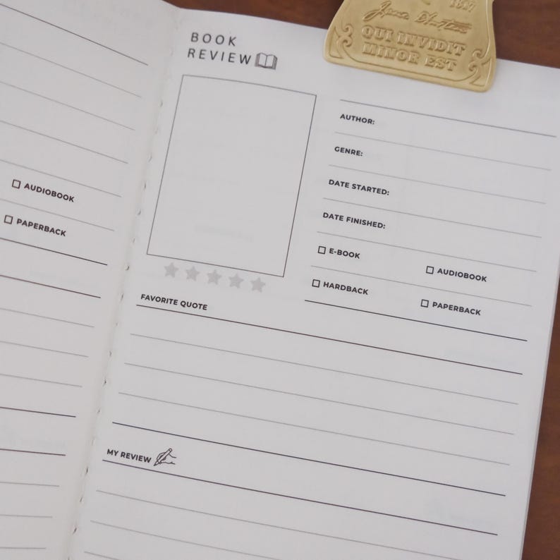 May include: Open notebook with book review template. The page includes sections for author, genre, and date. A gold-colored metal clip with the text "Qui invidit minor est" is attached to the page. The notebook is designed for book reviews.