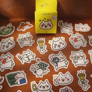 May include: A collection of cat-themed stickers and a yellow box. The stickers feature various cartoon cats with different expressions and holding objects like microphones and strawberries. The box reads "NIZMO-CARD" and "Cat's Mood".